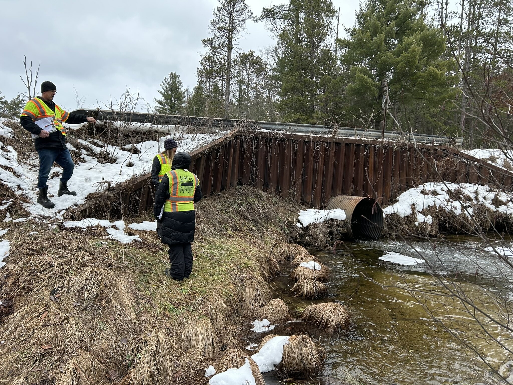 Before__Fife_Lake_Creek___County_Line_Road_(E_2_Road) Three people in high-visibility safety vests inspect an undersized metal culvert at Fife Lake Creek and County Line Road (E 2 Road), with snow-covered streambanks.