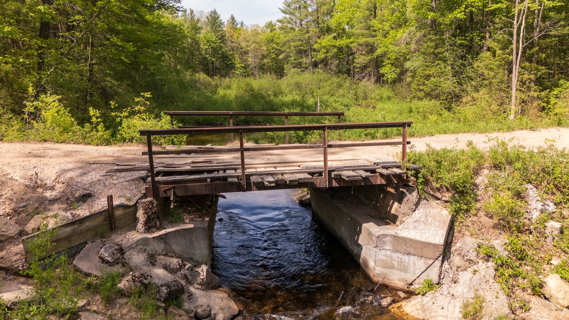 default Aging wooden bridge with uneven, deteriorating planks and metal railings spanning Fife Lake Outlet at Ramsay Road, surrounded by forested streambanks.
