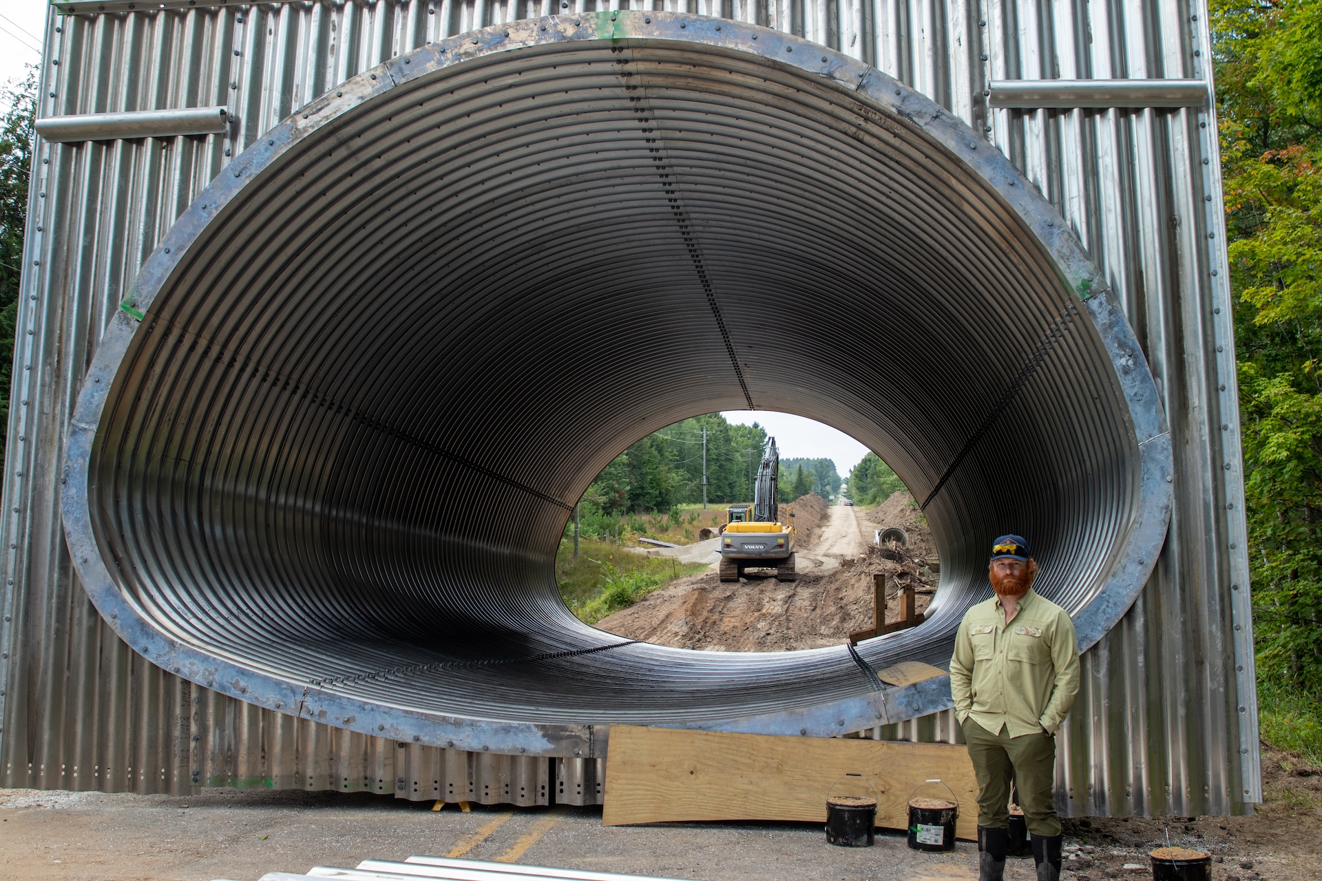 IMG_9613 CRA Project Manager Keenan Cooper stands beside a large corrugated metal stream crossing structure at Maple Creek and Sharon Road during construction in summer 2026, with an excavator visible through the opening.